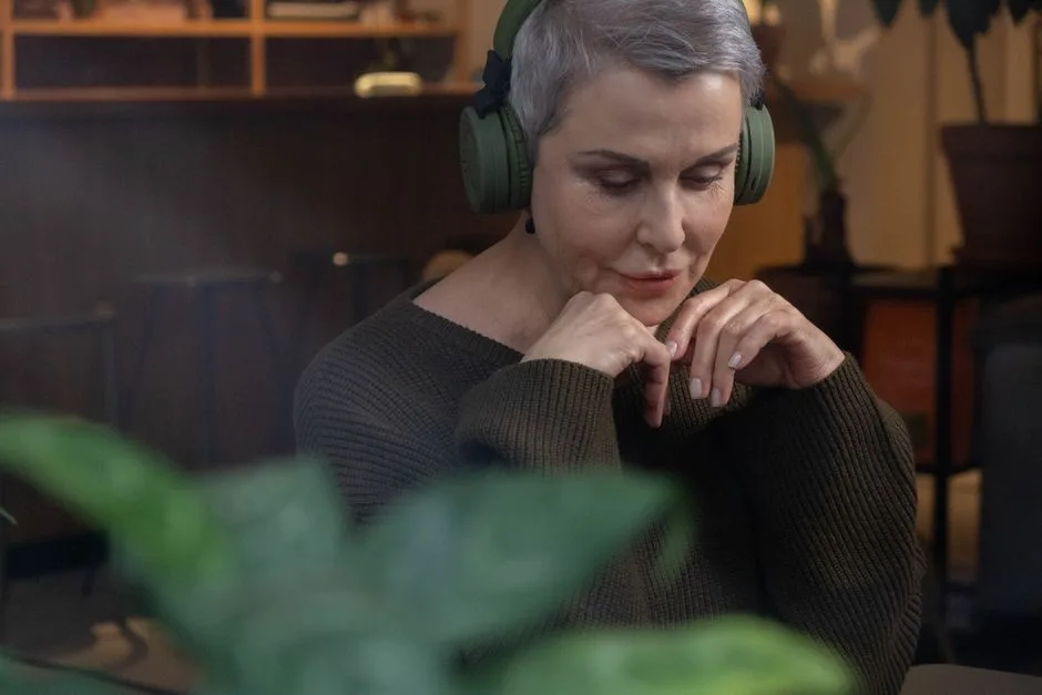 A senior woman wearing wireless headphones, relaxing indoors with a thoughtful expression.