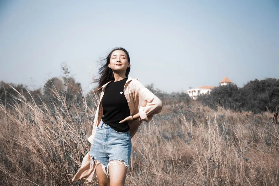 A young woman enjoying a sunny day in a field, eyes closed and hand on hip, exuding confidence.