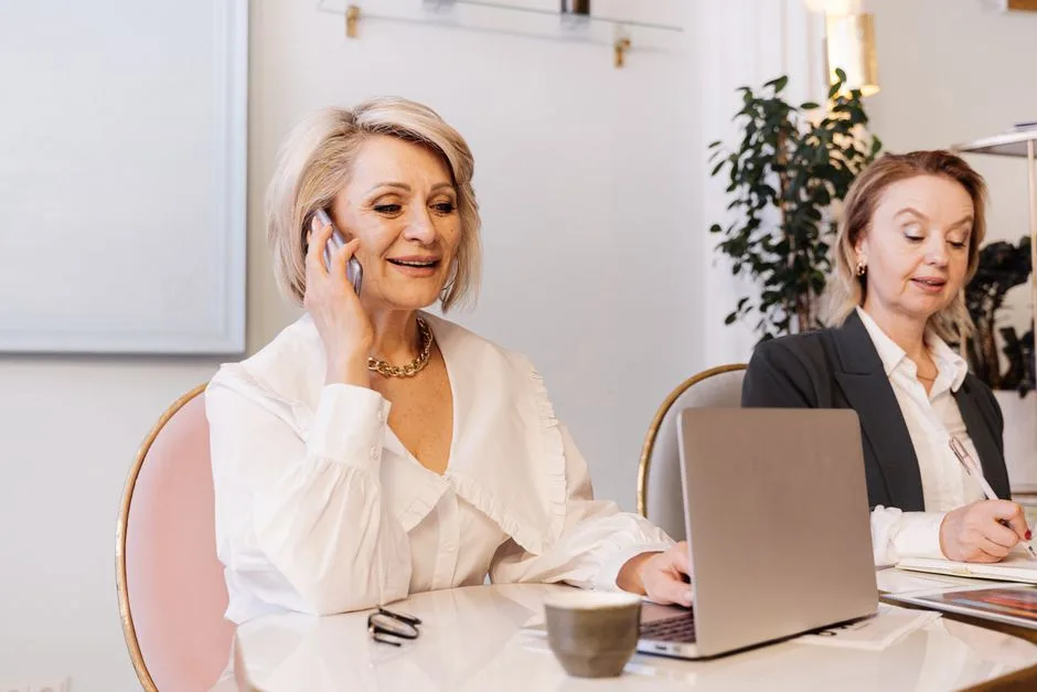 Two senior businesswomen engaged in work, using a phone and a laptop in a modern office.