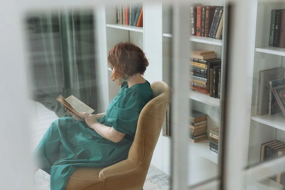 A woman enjoys reading in a comfortable chair surrounded by books in a home library.