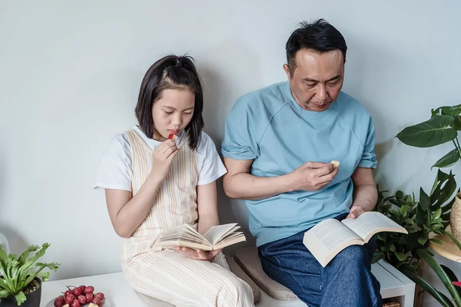 Father and daughter reading books together indoors, sharing a family moment with snacks and plants around.