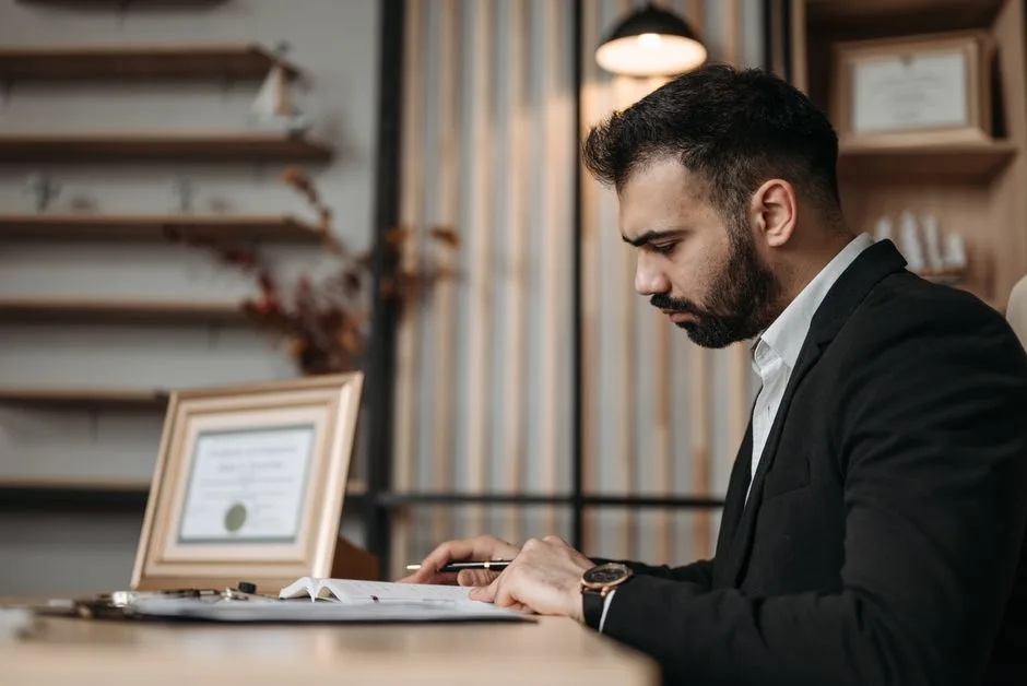 A professionally dressed man focused on writing at a desk within an office setting, suggesting a serious work environment.