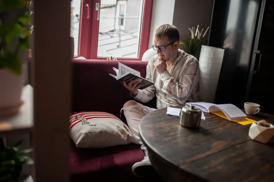 A man sits in a cozy room, reading a book by the window with a thoughtful expression.