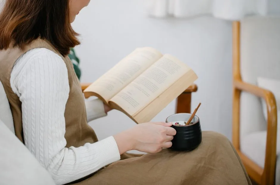 Woman sitting in an armchair reading a book and holding a cup of tea, in a peaceful indoor setting.