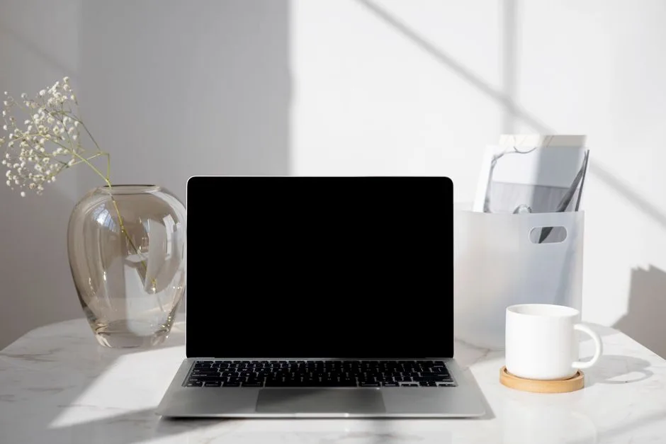 A clean and modern desk with a laptop, coffee cup, vase, and documents under bright daylight.