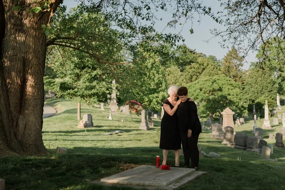 Two women stand together in a cemetery, paying their respects at a grave, surrounded by serene nature.