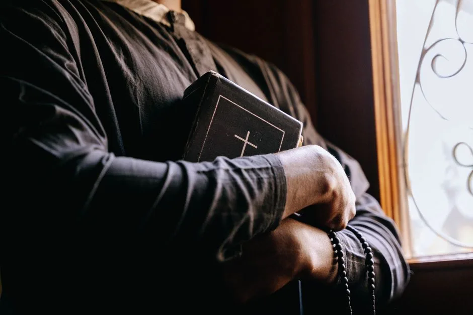 A priest holding a Bible by a sunlit window, symbolizing faith and spirituality.