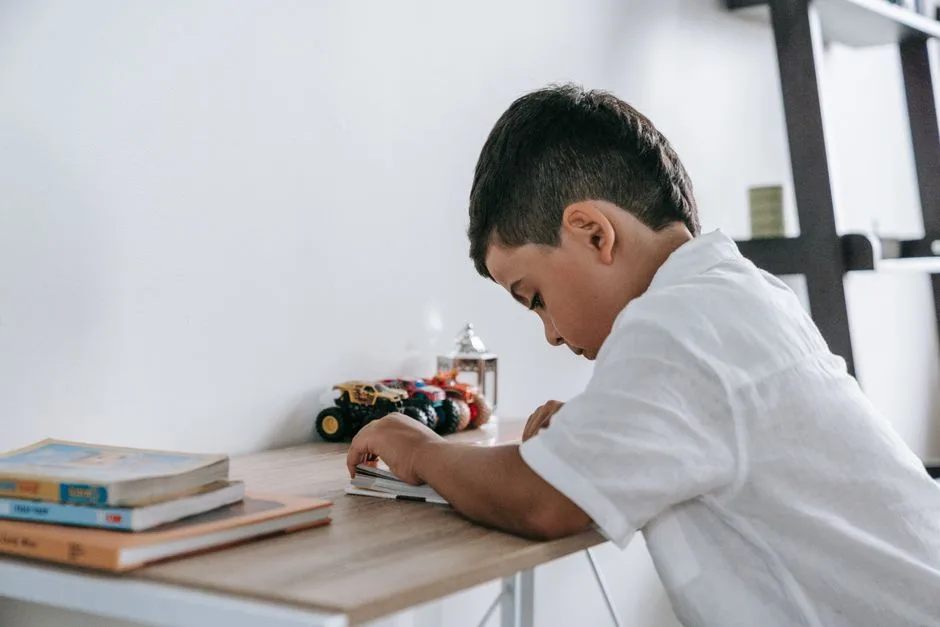 Child engrossed in reading at a desk with toy trucks in a bright room.