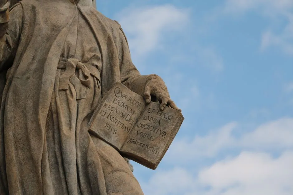 Close-up of a historical stone statue holding an open book against blue sky.