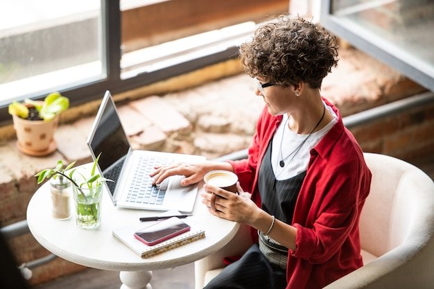 Pretty brunette female in smart casualwear having cappuccino while surfing in the net in front of laptop
