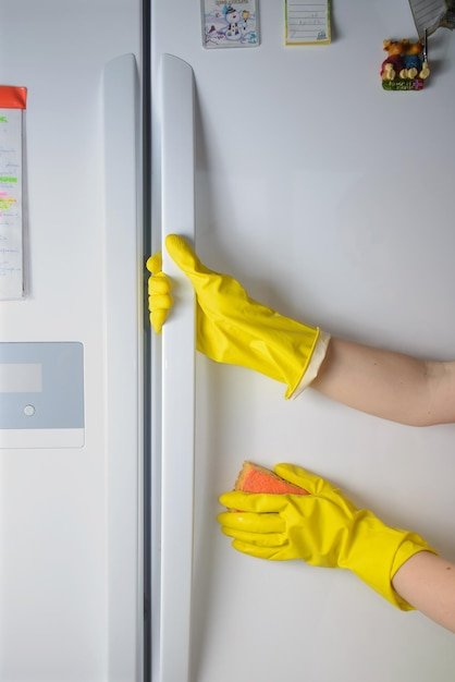 A Woman hand in yellow rubber protective glove cleaning white door of closed refrigerator with orange sponge