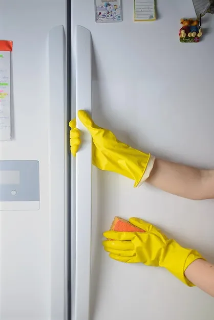 A Woman hand in yellow rubber protective glove cleaning white door of closed refrigerator with orange sponge