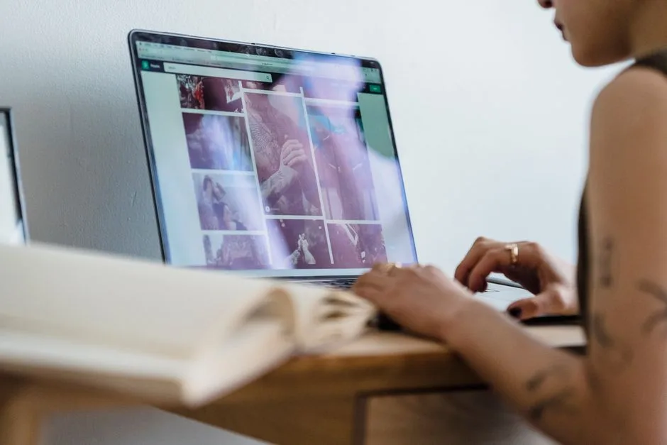 Crop unrecognizable female freelancer with tattoos sitting at table and browsing photos on laptop in room in daytime