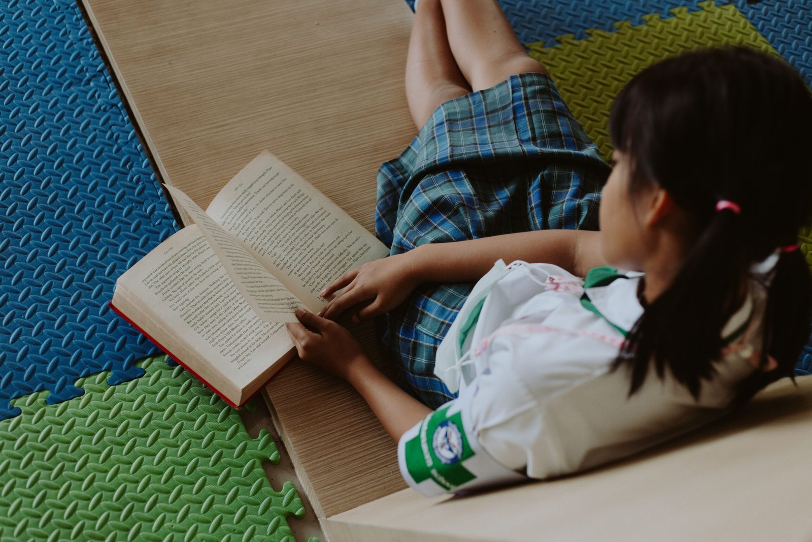 A young girl in a school uniform reading a book while sitting on a colorful mat indoors.