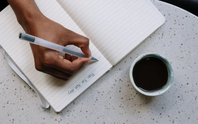 A person writes plans in a notebook beside a cup of coffee on a speckled table.