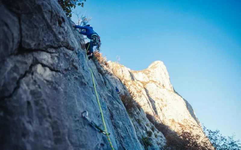 Adventurous rock climber ascending steep mountain face with safety gear in sunlight.