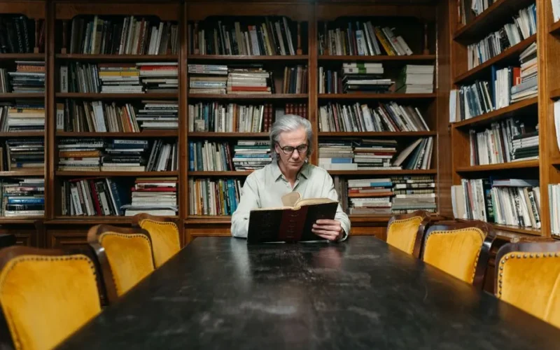 Senior man enjoying a book in a classic library with wooden bookshelves and a long table.