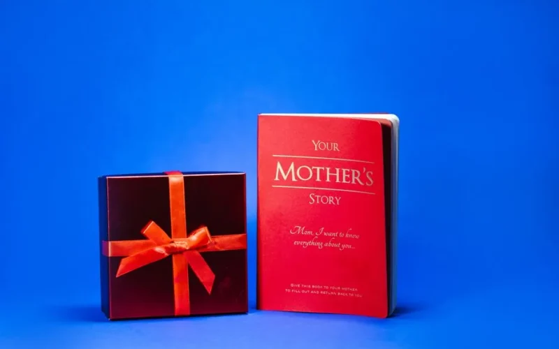 A wrapped gift box with red ribbon and a book titled 'Your Mother's Story' on a vibrant blue background, perfect for Mother's Day.
