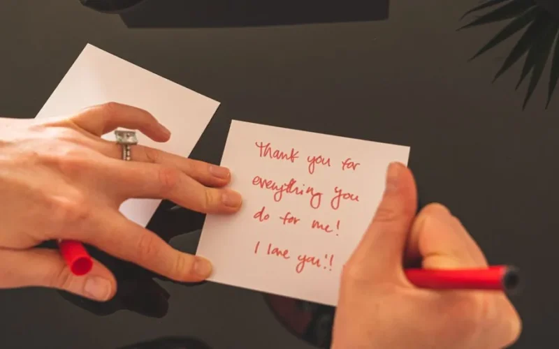 A person writing a heartfelt thank you note with a red pen on white paper.