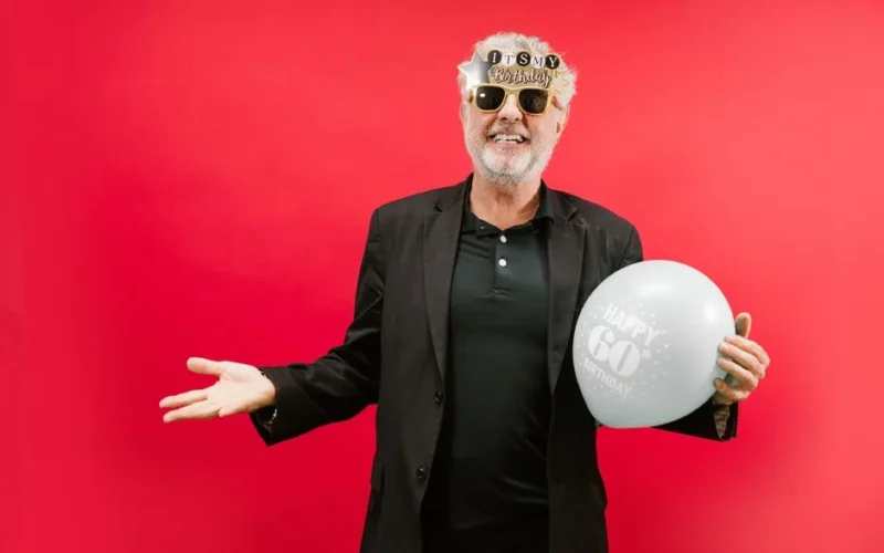 Happy senior man celebrating 60th birthday with sunglasses and balloon against a vibrant red backdrop.