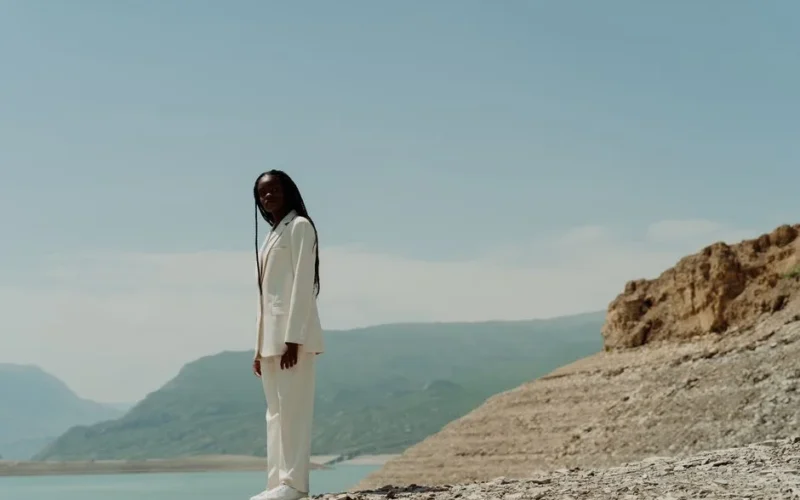 A woman in a white suit stands confidently against a scenic mountain backdrop under a clear blue sky.