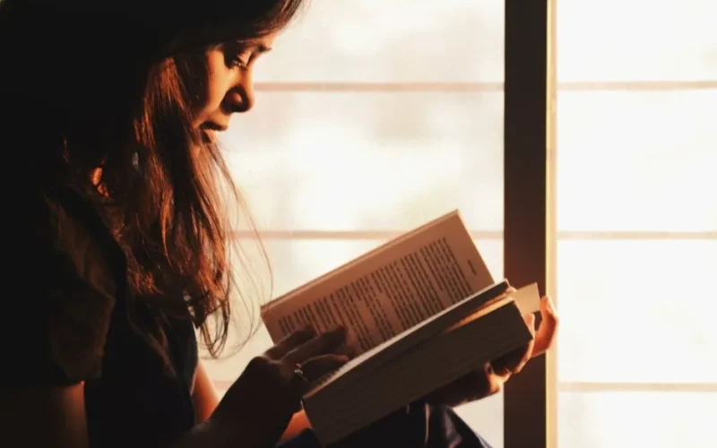 A woman engrossed in reading a book by a sunlit window creates a serene atmosphere.