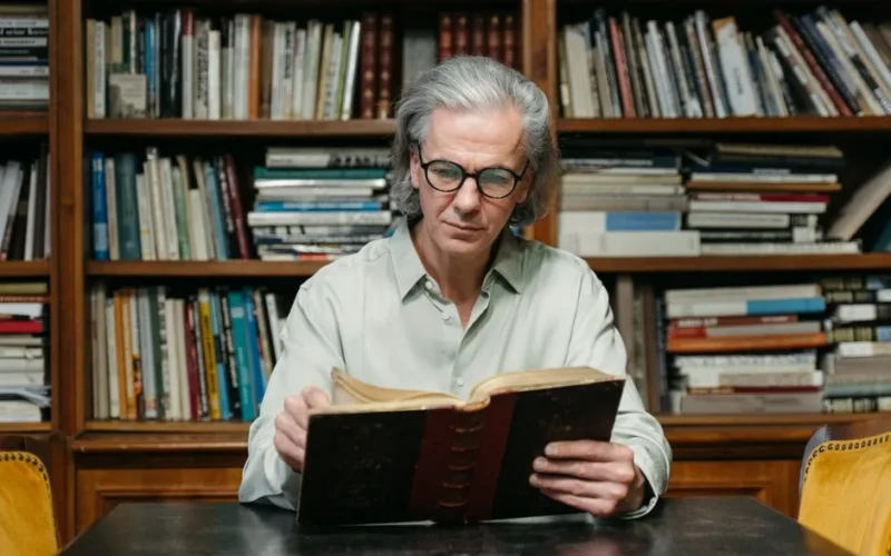 Senior man with eyeglasses reading a book in a library full of bookshelves.