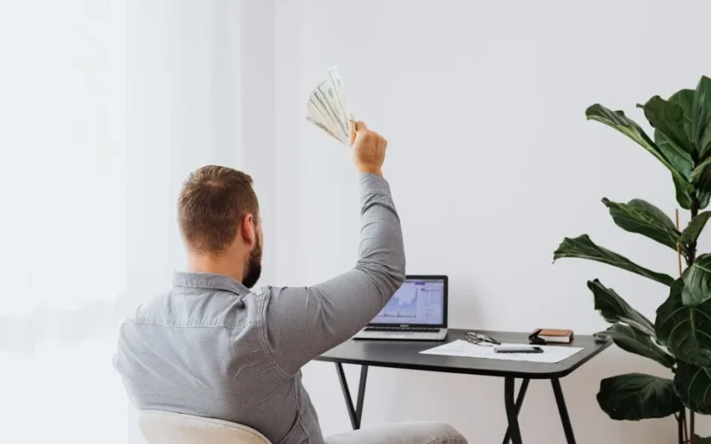 A man in a home office raises cash, symbolizing financial success and achievement.