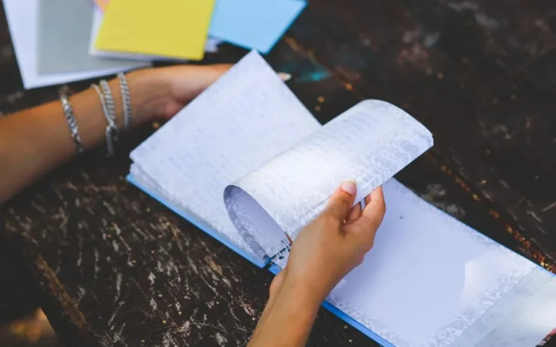 Close-up of hands flipping through a notebook on a worn wooden table. Outdoors setting, relaxed atmosphere.