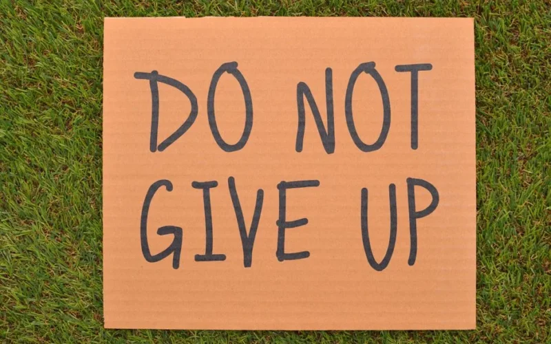 Cardboard sign reading 'Do Not Give Up' placed on green turf, symbolizing perseverance.