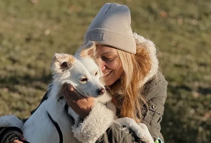 A woman in a beanie snuggles with her white mixed breed dog in a sunlit outdoor setting.