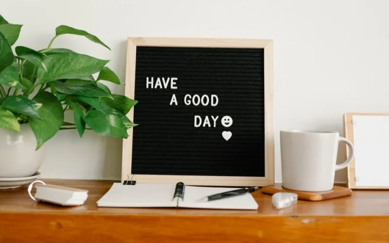 Various stationery and mug of hot beverage placed on shelf near board with Have A Good Day inscription and potted plant