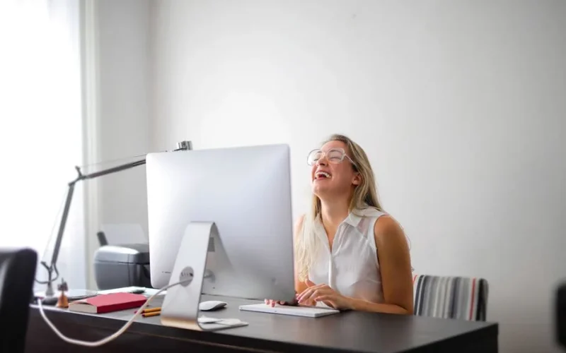 Office scene with a woman laughing while working at a modern desk, showcasing joy and productivity.