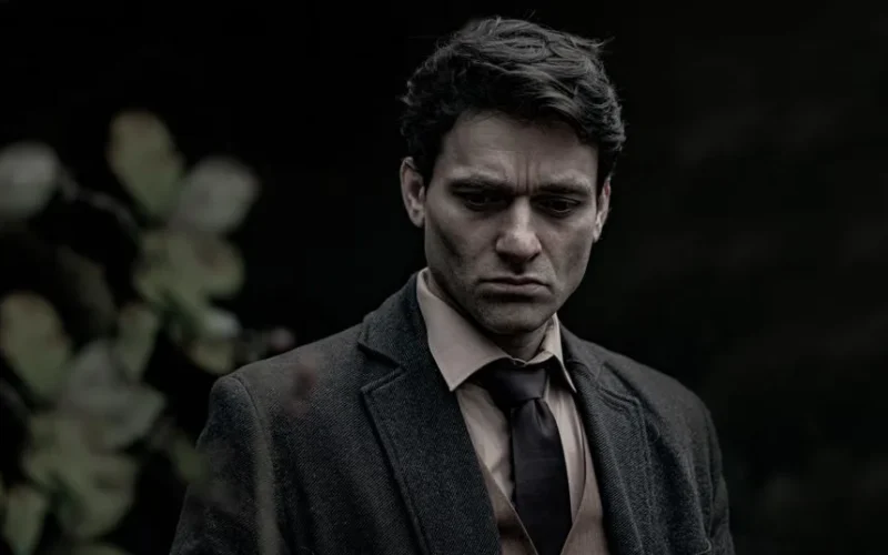 A pensive man stands outdoors in a dark suit, reflecting somberly.
