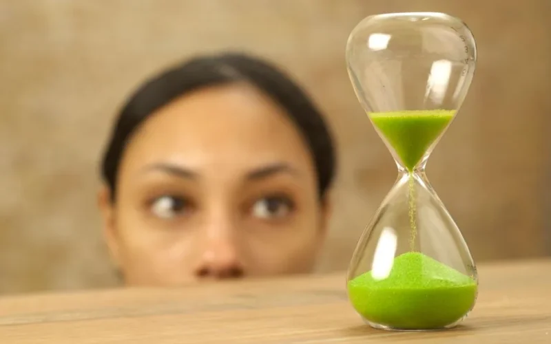 A focused woman looks at an hourglass with vibrant green sand. Concept of time and patience.