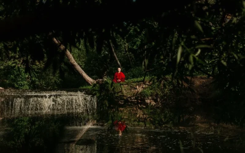 A serene scene of a Buddhist monk meditating near a tranquil waterfall, surrounded by lush greenery.