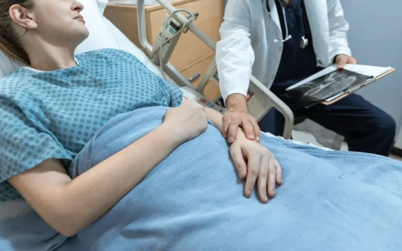 A doctor comforts a female patient in a hospital bed, holding her hand, with x-ray results.