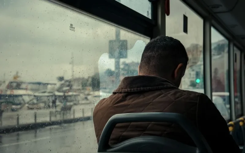 A solitary man on a bus gazes out at a rainy cityscape through a window.