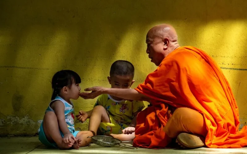 A Buddhist monk in orange robes feeds a young child against a yellow wall.