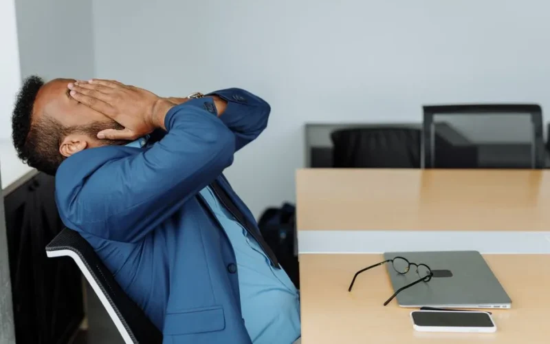 A stressed businessman in a suit covers his face while sitting at a desk with office tools.