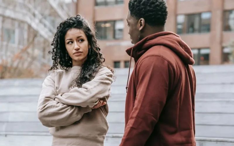 Unset ethnic woman with crossed arms and African American man looking at each other while having quarrel on street with building on blurred background