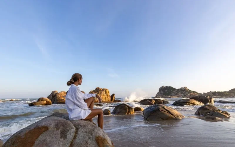 Woman in white sitting on rocks by the sea at sunrise, enjoying serene beachscape.