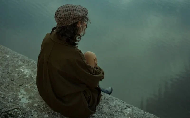 A contemplative man sits on a large rock surrounded by misty mountains, creating a serene and introspective scene.