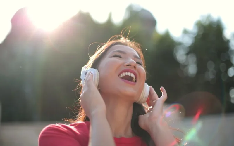 A cheerful woman in a red shirt enjoys music outdoors in the park with headphones.