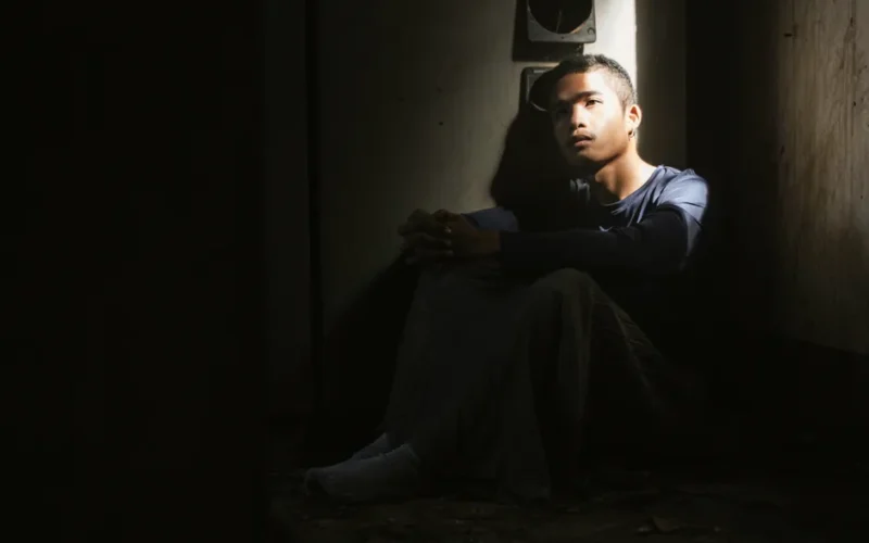 A young man sits alone in a dimly lit room, deep in thought.