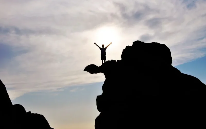 Silhouette of a person celebrating on a mountain peak against a dramatic sky, symbolizing freedom and success.