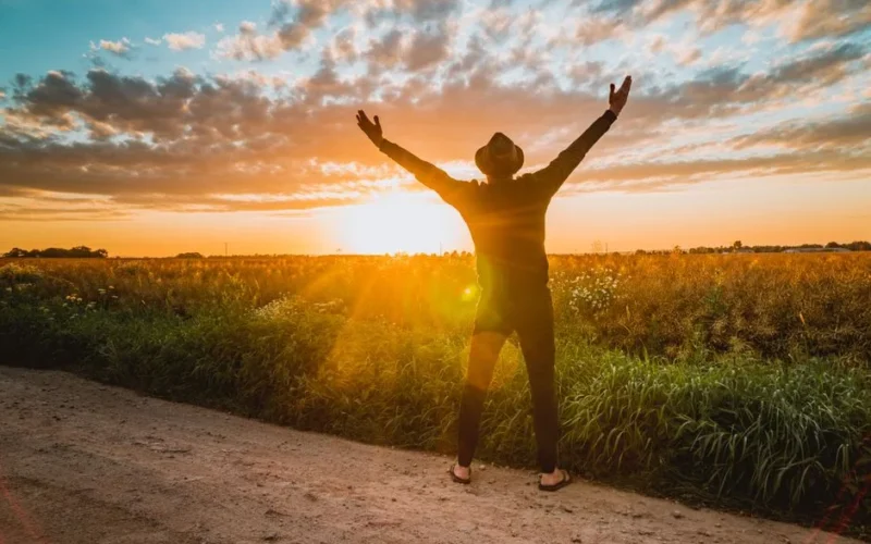 A man raises arms in freedom at sunrise in a rural field, expressing joy and connection with nature.