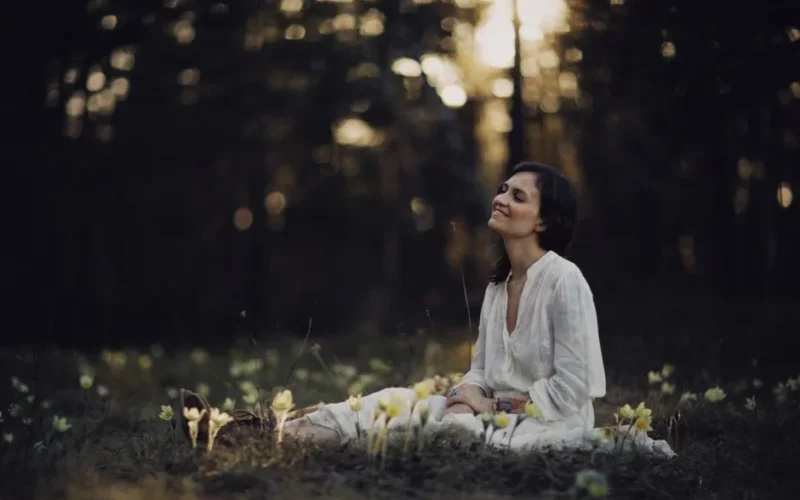 A woman sits peacefully in a sunlit forest clearing surrounded by flowers.