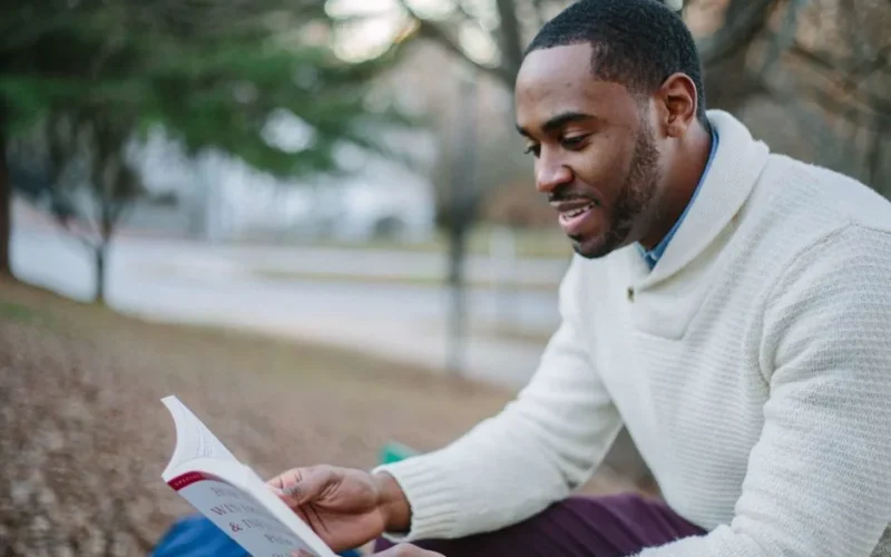 A young man enjoys reading in a serene park setting during the day.