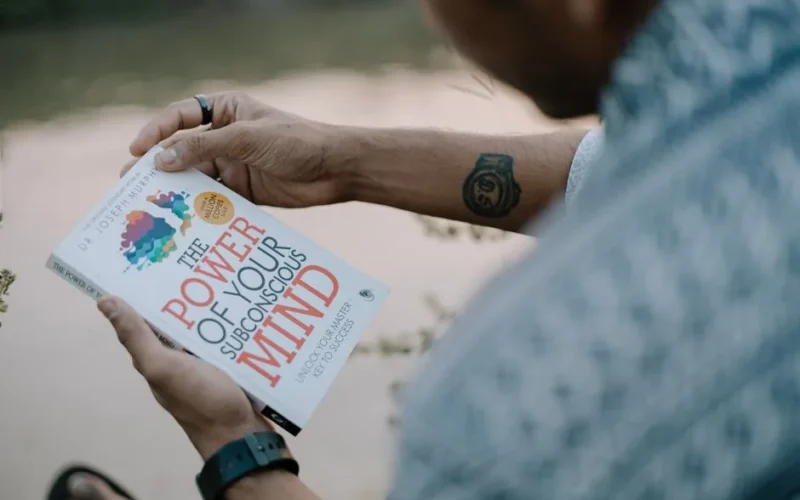 A man reads 'The Power of Your Subconscious Mind' by a riverside, showcasing relaxation and reflection.